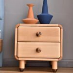 Amyzel rounded solid wood kids nightstand with two drawers and checkerboard knobs, styled with a modern orange and blue vase on top against a gray wall.