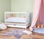 Styled nursery featuring the Amyzel white wooden cot bed with an integrated under-bed drawer, tucked under a flowing pink fabric canopy next to a large teddy bear.
