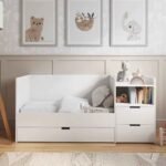 White wooden convertible crib shown transformed into a child's daybed with front slats removed, styled with plush pillows and an attached side storage drawer unit.