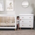Lifestyle nursery setting with the white 3-drawer dresser placed next to a wooden slat baby crib, featuring a toddler standing between them.