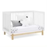 White convertible baby crib transformed into a toddler daybed styled with grey throw pillows and a polka-dot fitted mattress sheet.