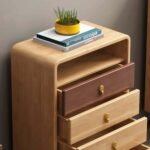 Close-up of a Nordic light wood storage cabinet with smooth curved corners, an open top cubby shelf holding books and a potted plant, and pulled-open drawers with gold knobs.