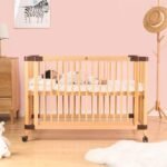 Natural solid wood slatted baby cot with wheels, featuring a baby sleeping comfortably inside a soft pink nursery setting.
