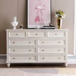 Frontal view of an antique off-white 9-drawer wide dresser with gold hardware placed in a styled living room setting.