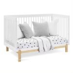 White convertible baby crib transformed into a toddler daybed styled with grey throw pillows and a polka-dot fitted mattress sheet.