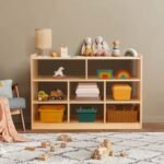 Natural wood low toy storage unit styled in a serene nursery room setup with stuffed plush animals on top, books, wooden vehicles, and woven bins.