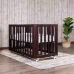 Dark espresso walnut wooden mobile baby crib styled on a geometric rug in a modern nursery with birch tree wallpaper and a potted green houseplant.