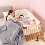 Natural finish solid wood co-sleeper crib aligned directly against an adult bed, featuring a mother lying down and checking on her sleeping baby.