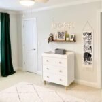 Lifestyle image of the white 3-drawer chest of drawers set up in a bright nursery room with wall decor, green curtains, and a plush rug.