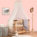 Natural wood baby crib styled with an elegant, full-length white sheer mosquito net canopy suspended from overhead in a nursery.