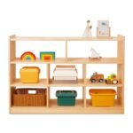 Modern natural wood low Montessori bookshelf with 8 asymmetric storage compartments filled with wooden toys, rainbow stackers, and colorful storage bins on a white background.