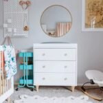 Styled nursery setup showcasing the white 3-drawer dresser flanked by a teal 3-tier rolling cart, a round wall mirror, and a baby bouncer chair.
