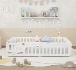 Pure white wooden kids floor bed with a full slatted safety enclosure and a small gate, styled with pastel plush pillows on a large round white rug.