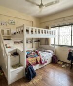 Modern white solid wood twin bunk bed with an integrated left-side storage staircase featuring drawer steps, set in a brightly lit children's bedroom next to a large window.