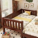 Rich mahogany finish wooden toddler co-sleeper bed extension attached to a larger bed, featuring high slatted safety rails, elephant-printed sheets, and a yellow blanket panel.
