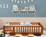Warm honey oak finish children's floor bed featuring a secure slatted safety rail with a small access gate, arranged on a colorful polka dot rug against a slate blue wall.