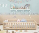Light natural pine wood toddler floor bed with a protective slatted frame enclosure, styled in a cozy nursery room with bunting flags, string lights, and a tree-patterned wall border.