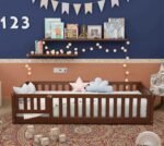 Amyzel premium dark walnut finish wooden toddler floor bed featuring a full slatted safety guardrail enclosure with a small latch gate, adorned with star and cloud pillows on a traditional red patterned rug.
