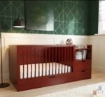 Rich dark mahogany finish multi-functional wooden baby crib, featuring a built-in side storage unit with drawers and open shelves, complete with a large under-bed drawer against a dark green geometric accent wall.