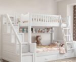 Pure white painted wooden kids bunk bed with a safety guardrail on the upper bunk, an integrated side storage staircase, and a front ladder, styled with a plush bear toy.