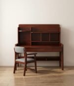 A solid wood study desk with a rich mahogany finish, shown against a plain light wall on wood flooring. The desk includes an attached upper hutch with open shelving compartments, two sleek pull-out drawers, and an additional lower storage shelf near the base. A matching mahogany-stained wooden chair with light grey cushioned seating and backrest sits in front of the desk.