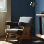 Charcoal grey fabric lounge chair with warm timber V-frame legs set against a deep navy feature wall with a brass floor lamp.
