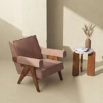 Mid-century modern accent chair in dusty rose upholstery, featuring angled V-shaped solid wood legs and padded block armrests beside a stone-top tripod side table.