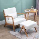 Modern white fabric lounge armchair with natural wood frame, matching ottoman footstool, and round wooden end table