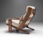 Side back profile view of a mid-century modern lounge chair featuring a solid dark wood angular frame and thick beige linen cushions.