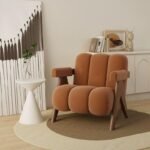 Rust terracotta channel-upholstered accent chair with wooden V-frame legs set on a beige circular rug next to a white side table.