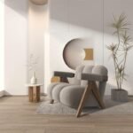 Light grey modern channel-tufted plush armchair with solid wooden legs in a sunlit interior with minimalist wall arches.