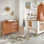 Scandinavian style baby nursery room layout, Amyzel dino shaped kids accent rug, wooden changing table chest of drawers, white infant crib with warm canopy.