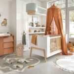 White wooden infant cot bed with orange fabric canopy drape, Amyzel dinosaur shaped plush nursery floor mat, child friendly bedroom storage shelves and dinosaur basket.