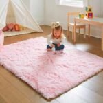 Amyzel soft pastel pink fluffy shag rug for girls bedroom, high pile plush pink nursery area carpet with a toddler playing safely on the floor near a wooden table set.